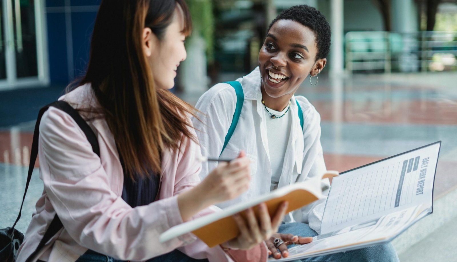 Happy young multiracial women in casual clothes sitting outdoors with notepad and papers in daytime while looking at each other
