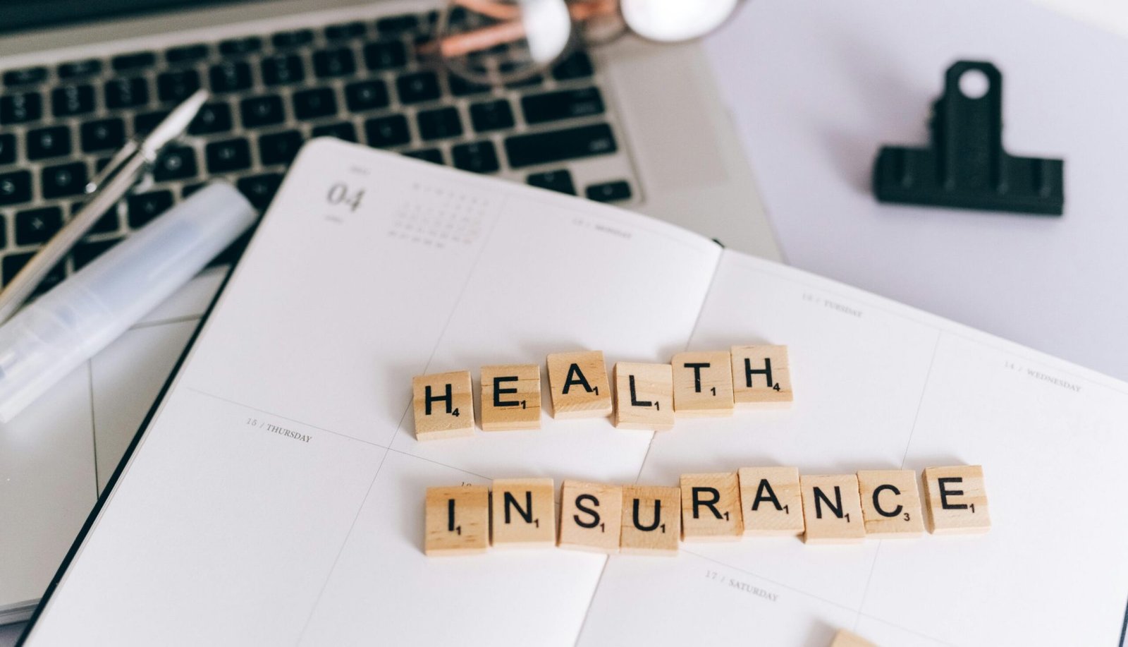 Close-up of Scrabble tiles spelling 'Health Insurance' on a planner with a laptop.